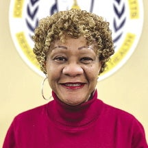 A woman with blonde curly hair wears a red turtleneck and silver hoop earrings.