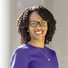 A woman with glasses wears a purple top and pearl earrings.