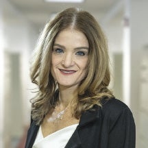 A woman with blonde hair wears a black jacket and white blouse.