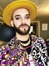A person wears a yellow hat, a purple, black, yellow, white, and red patterned jacket, and a gold necklace.