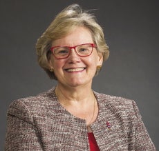A woman with short blonde hair wears red glasses and a tweed jacket.