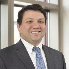 A man in a grey suit jacket, white button down, and blue patterned tie