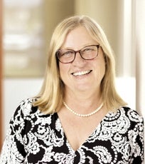 A woman with straight blonde hair wears large glasses, a pearl necklace, and a black and white paisley blouse.