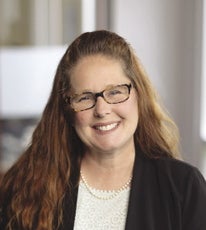 A woman with long brown hair wears a glasses, a black suit jacket, pearl necklace, and white lace blouse.