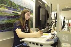 A woman with long brown hair wears blue scrubs standing in front of a painting of a hilly landscape, writing with a pink pen in front of a computer on a rolling cart. 