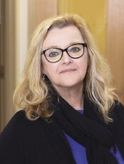 A woman with blonde hair wears large glasses, a black cardigan, purple sweater, and black knit scarf.