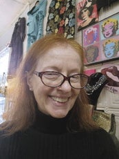 A woman with red shoulder length hair, a black turtle neck, and large glasses smiles in front of a wall with clothes hung up against it.