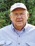 A man wears a white baseball cap, glasses, and a light blue button down shirt.