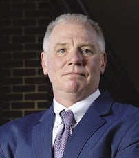 A man with white hair wears a blue suit with a tie.