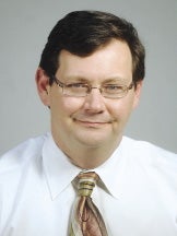 A man with brown hair and glasses wears a white button down shirt and a purple, blue, and green striped tie.