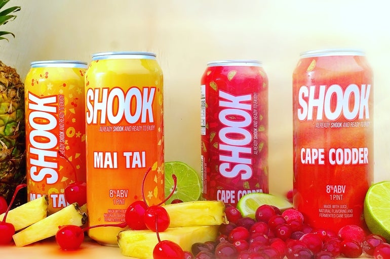 Orange, yellow and red cans of the alcoholic beverage SHOOK backdrop some slices of pineapple, cherries, limes and cranberries.