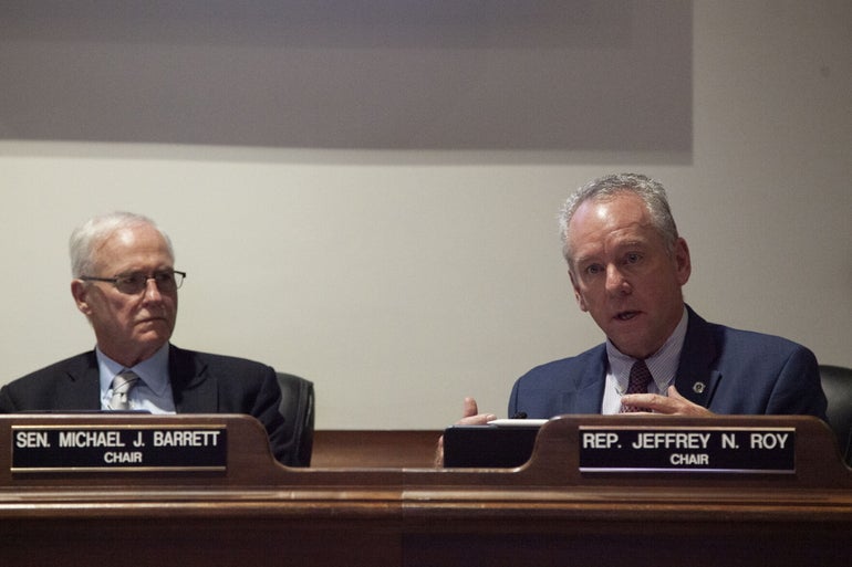 Two men in suits sit behind a desk with name placards in front