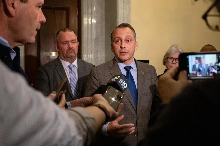 A man in a suit and blue tie talking in front of a group of reporters