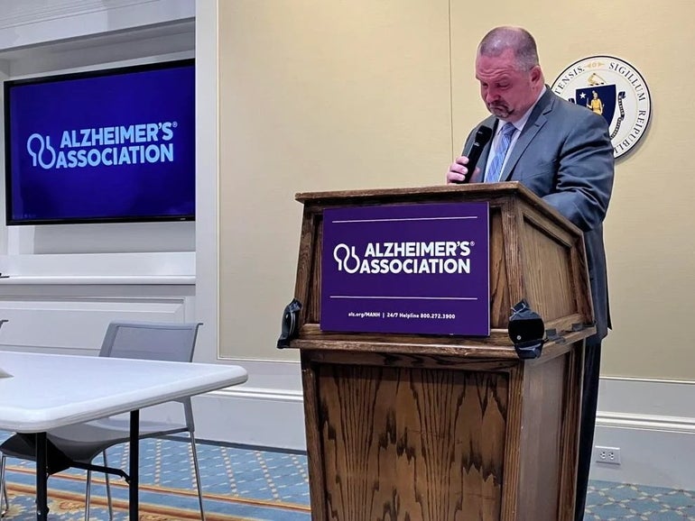 A man gets emotional while holding a microphone and standing behind a podium with the words "Alzheimer's Association".