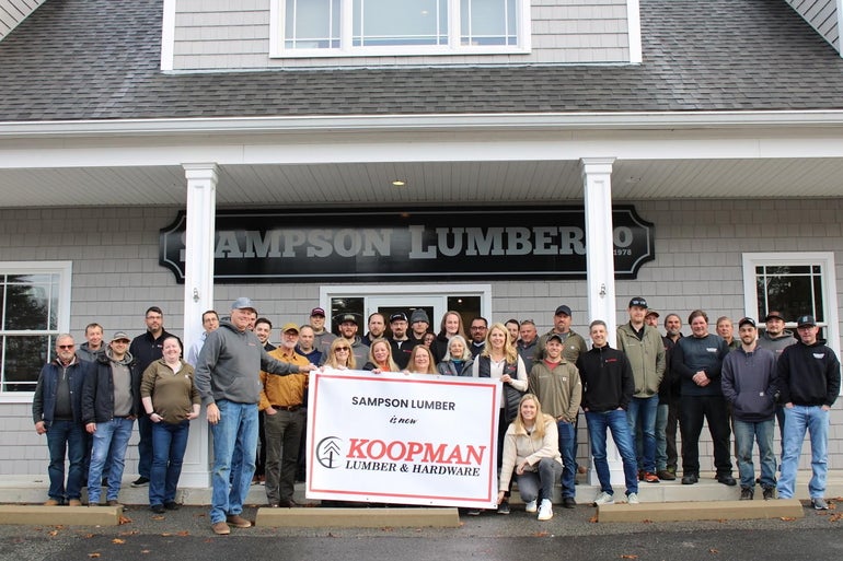 A goup of peiple stand outside a building with grey siding holding a white sign with red and black lettering.