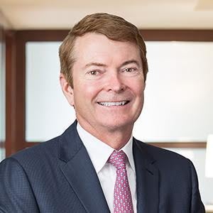 A man with light brown hair wears a dark blue suit, white button down, and red patterned tie.