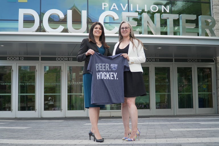 Two women in heels stand holding a t-shirt that says "Beer. Hockey." in front of an arena entrance