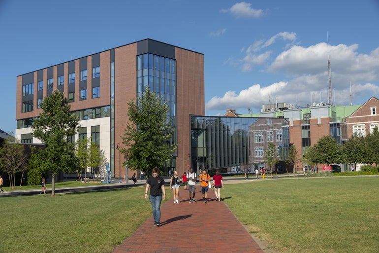 Students walk through a quad on a college campus with academic buildings behind them