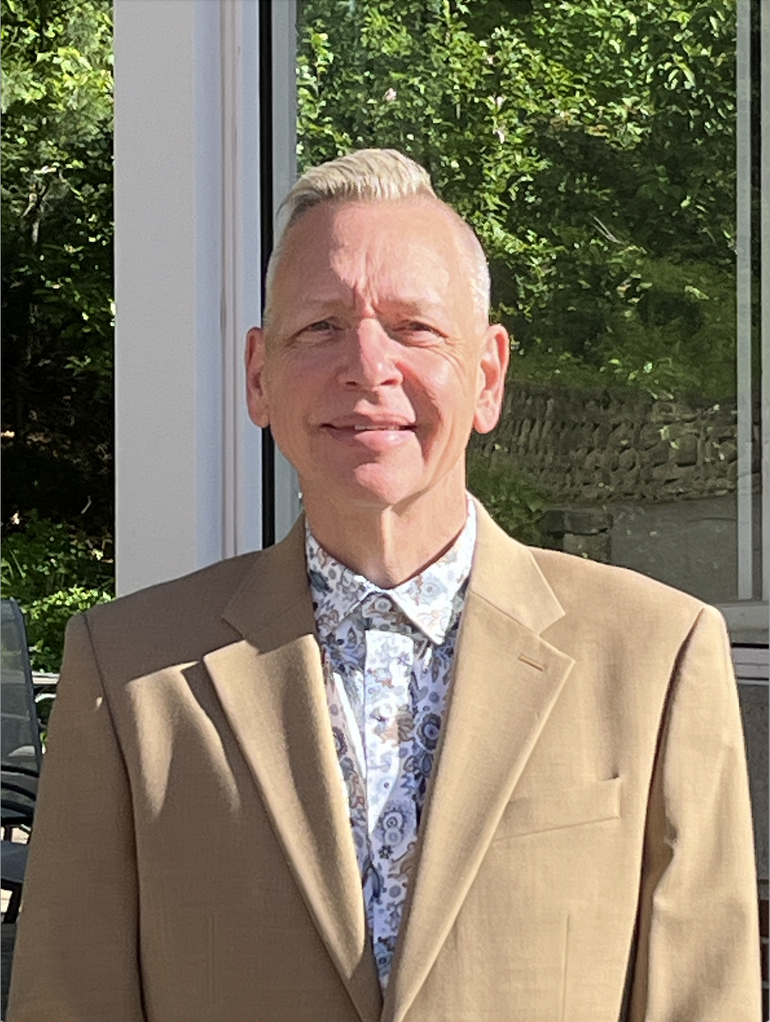 A man with blonde hair wears a light tan suit jacket and patterned button down.