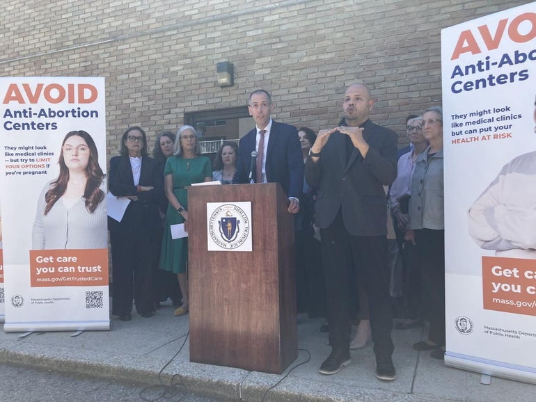 A man stands behind a wooden podium on a concrete sidewalk with 9 others around him in between signs reading "AVOID Anti-Abortion Centers".