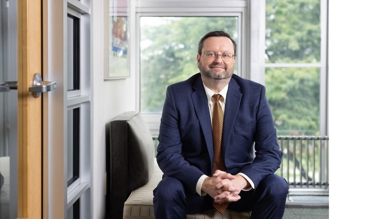 A man with glasses wears a dark blue suit and sits on a couch in front of windows with his hands held together.