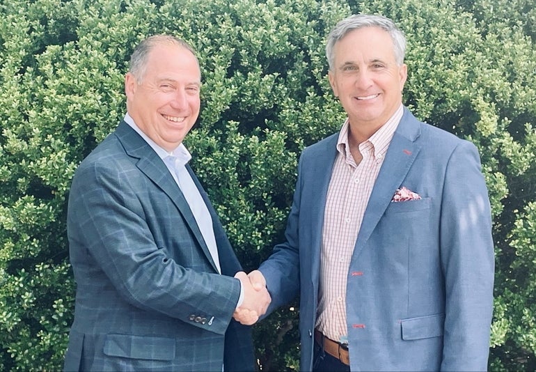 A man in a plaid grey/green suit jacket and light blue button down shakes hands with a man in a light blue suit jacket and red and white checkered button down in front of a large green shrub.