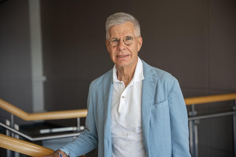 A man with white/grey hair and tortoise colored glass wears a light blue jacket and white polo with his hand on a wooden stair railing.