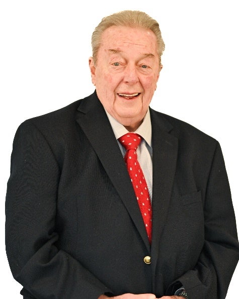 A man wears a black suit jacket, tan button up shirt, and red tie. 