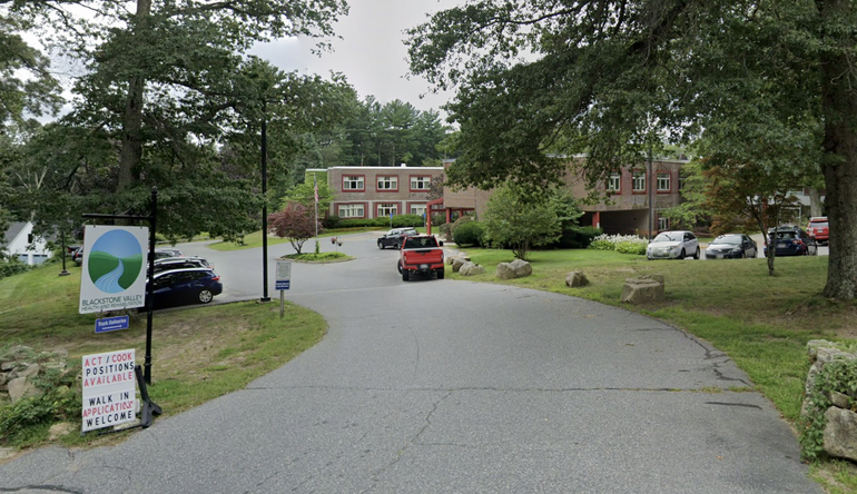 A paved driveway leads to a brick building partially covered by trees.