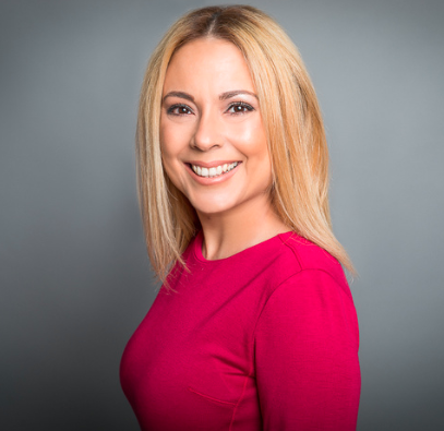 A woman with long blond hair wears a  red top in front of a grey background.