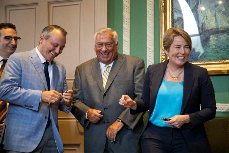 A woman in a suit and two men in a suit laugh while holding pens