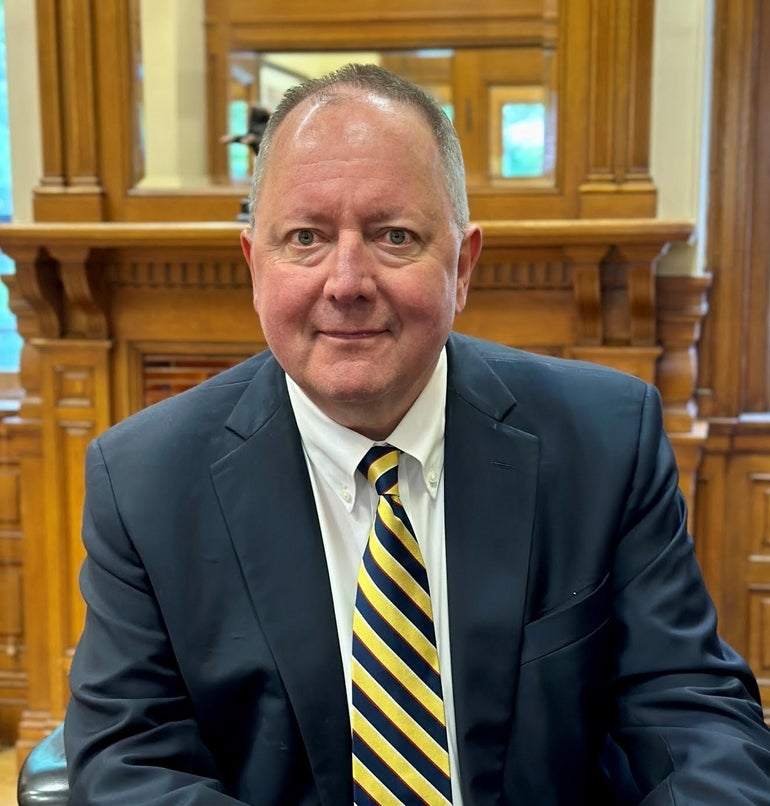 A man wears a dark blue suit jacket, white button down, and dark blue and yellow stripped tie sitting in front of a wooden mantlepiece.