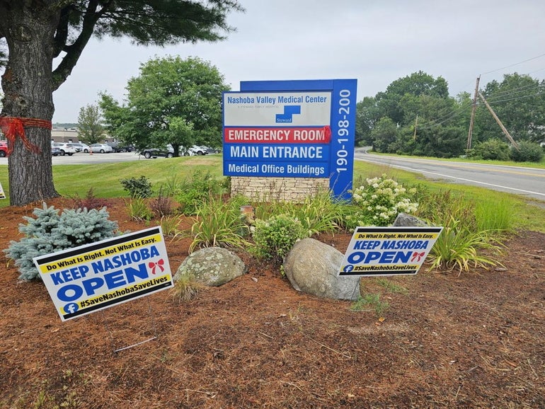 A sign reading "emergency room" and "main entrance" sits behind signs reading "keep nashoba open" on a mulch ground with a street running a long the right hand side.