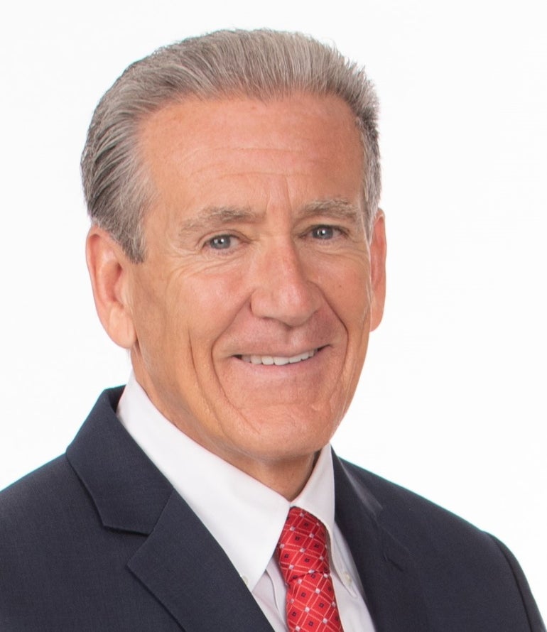 A man with grey hair wears a dark blue suit jacket, white button down, and red patterned tie.