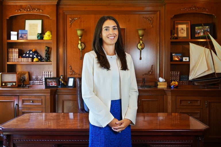 A woman with long dark brown hair wears a white top, a white blazer, and a blue skirt while standing in front of a wooden desk.