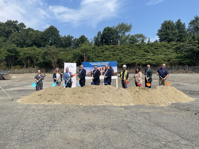 Officials gather for a groundbreaking ceremony behind a dirt pile with shovels in hand