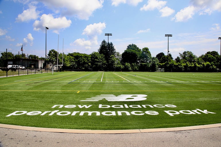 A turf athletic field with trees and light poles behind it