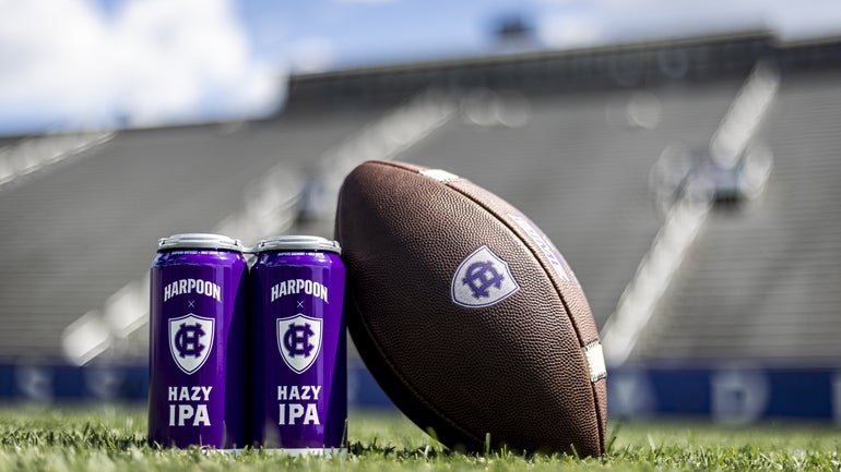 A four-pack of beer with a football leaning against it, a stadium behind both