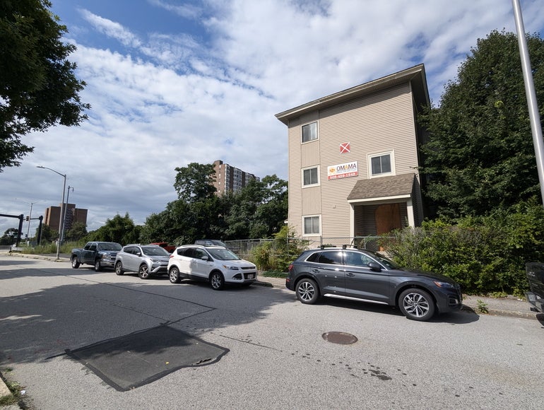 A vacant three-story building with fencing and overgrown shrubs around it and four cars parked in front