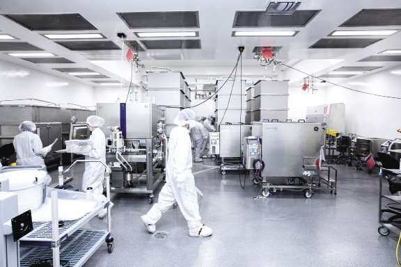 Workers wear white jumpsuits, face masks, and hairnets while working within a manufacturing facility with white walls.