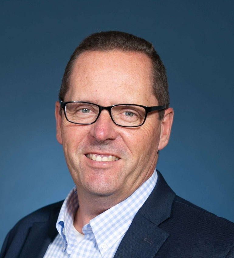 A man wears glasses, a white and light blue checkered button down, and dark blue suit jacket.