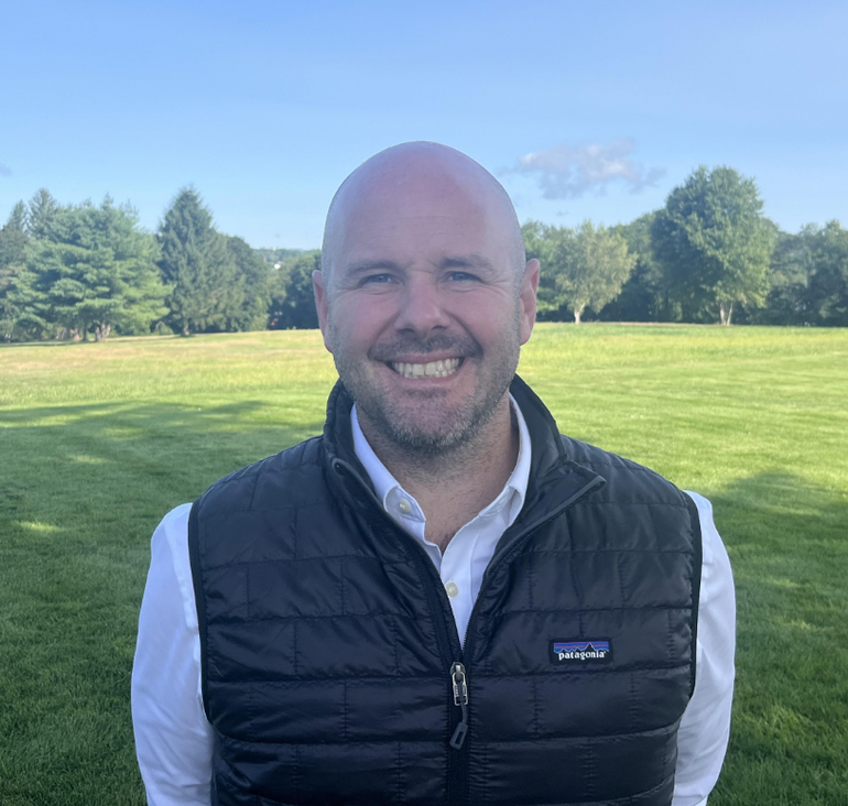 A man wears a white button down and black puffer vest, smiling in a field.