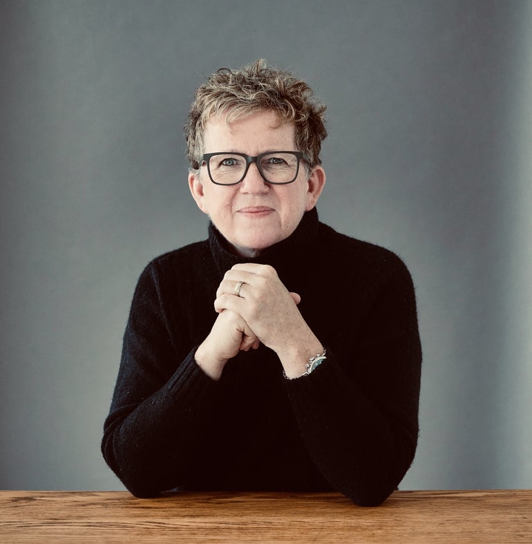 A woman with short grey hair wears large black glasses and a black turtleneck sweater sitting at a wooden table with her elbows on the table and hands held together in front of her chest.