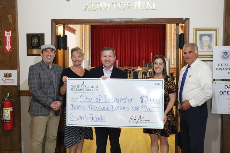 A group of people hold a large check in front of an indoor entrance to an auditorium.