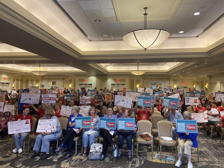 People sit on rows of chairs holding protest signs inside a banquet hall.