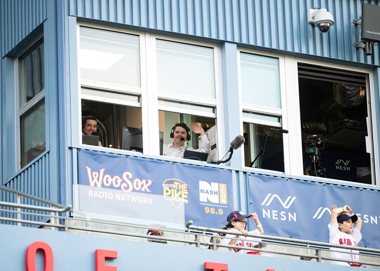 A man waves to the camera through the window of a baseball press box