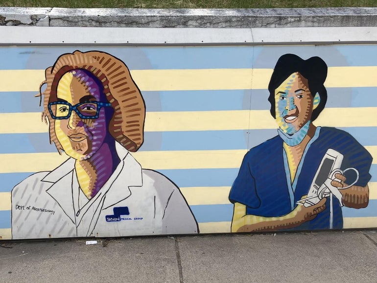 A mural of a doctor in a white lab coat and a nurse in blue scrubs against a stripped blue and yellow background.