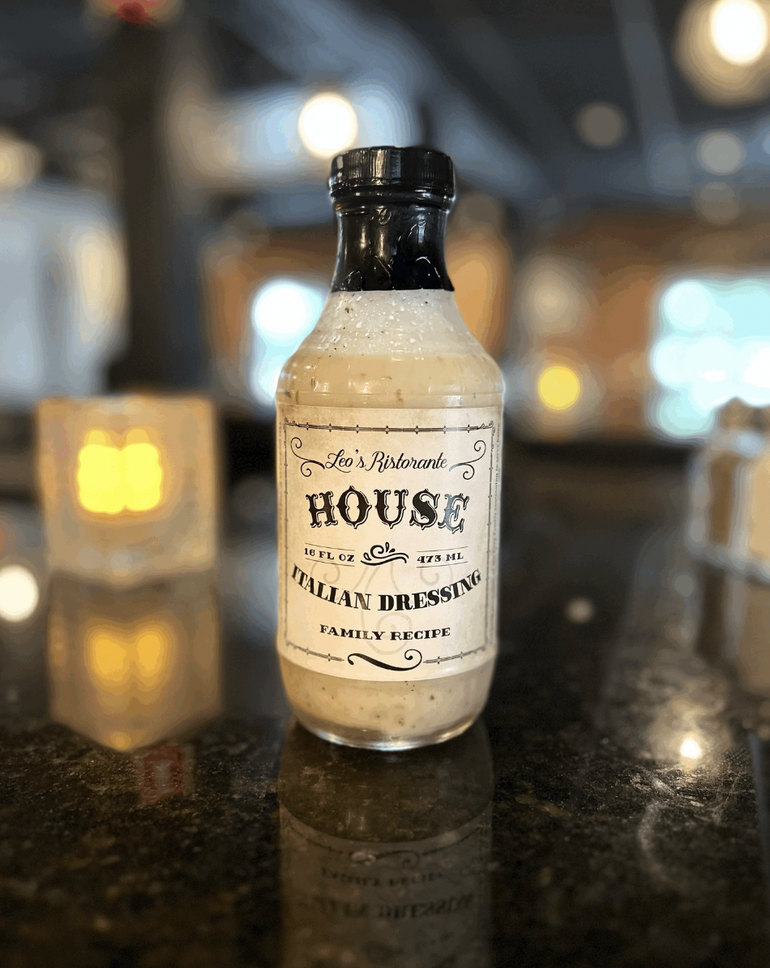 A salad dressing bottle with white dressing inside and a black top sits on a black countertop with a candle in soft focus behind it.