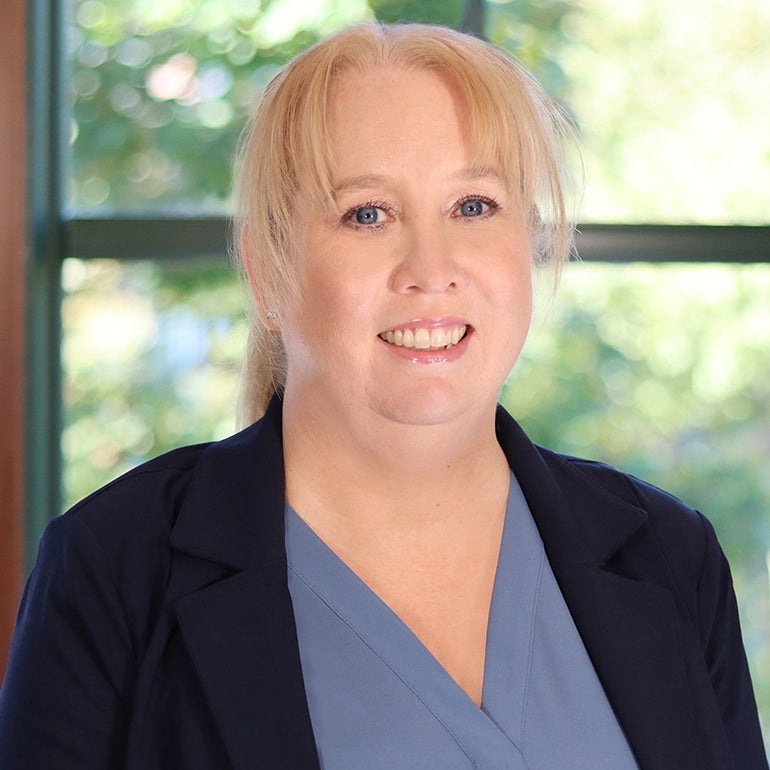 Headshot of a woman with blonde hair tied back with bangs wears a black suit jacket and blue blouse