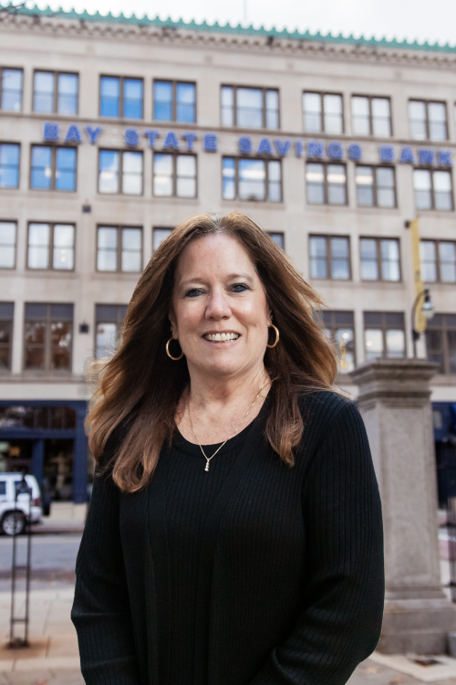 A woman with long brown hair wears a black blouse and gold hoops while standing in front of a building with "Bay State Savings Bank" on its outer wall.